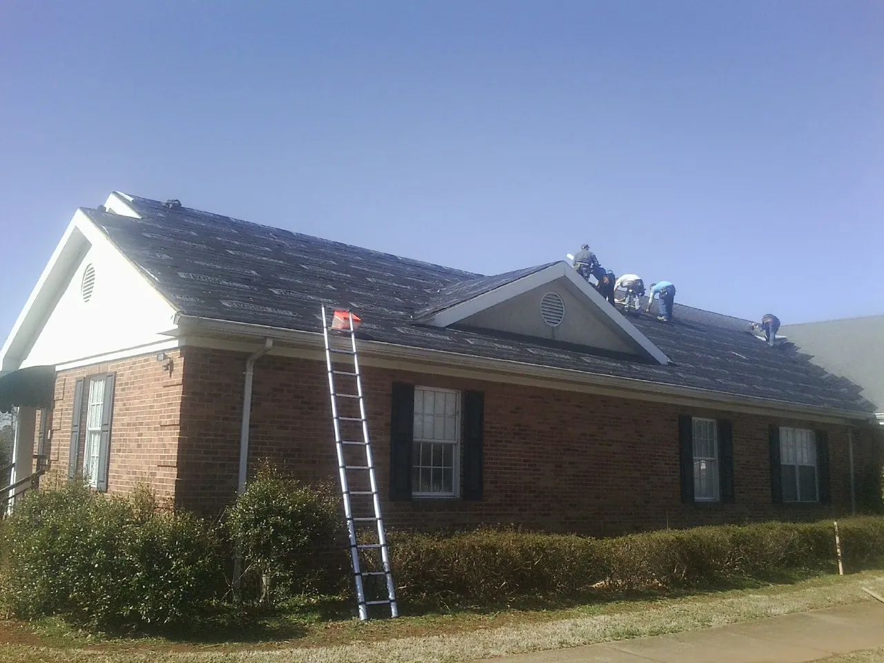 Crew tearing shingles off a brick ranch roof with a ladder set — Emergency Roof Repair work by our crew in Gonvick