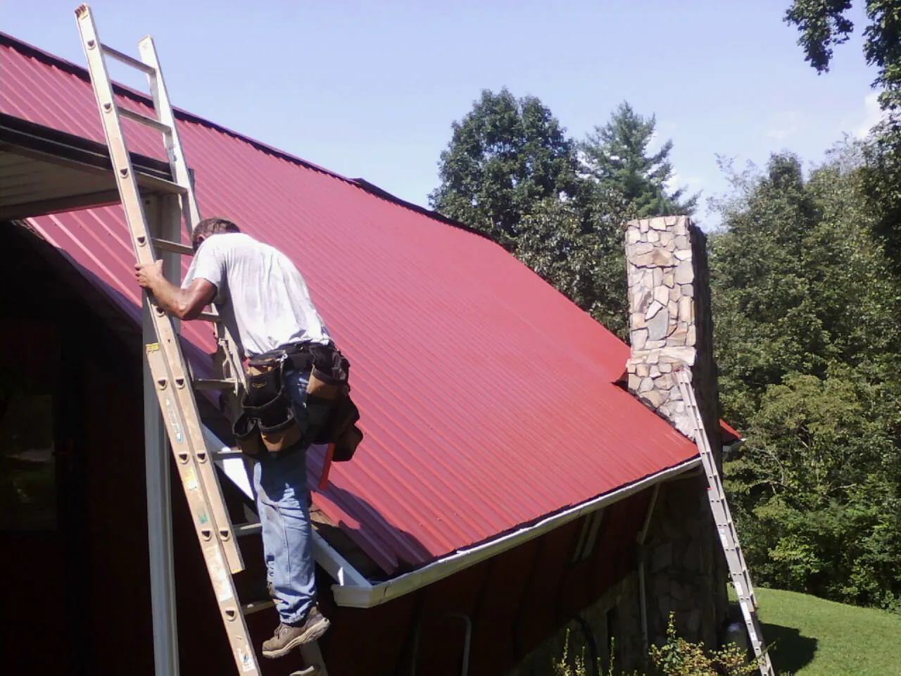 Roofer on a ladder beside a red metal roof and stone chimney — Soffit & Fascia Repair by Roofing Brothers in Gonvick